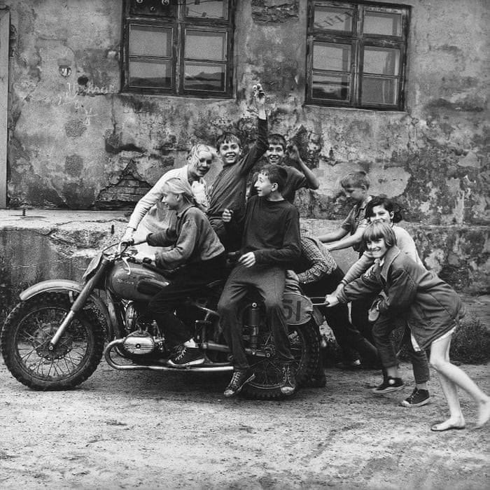 Photograph by Sonta. Children pose and crowd around an old motorcycle against the facade of a building.