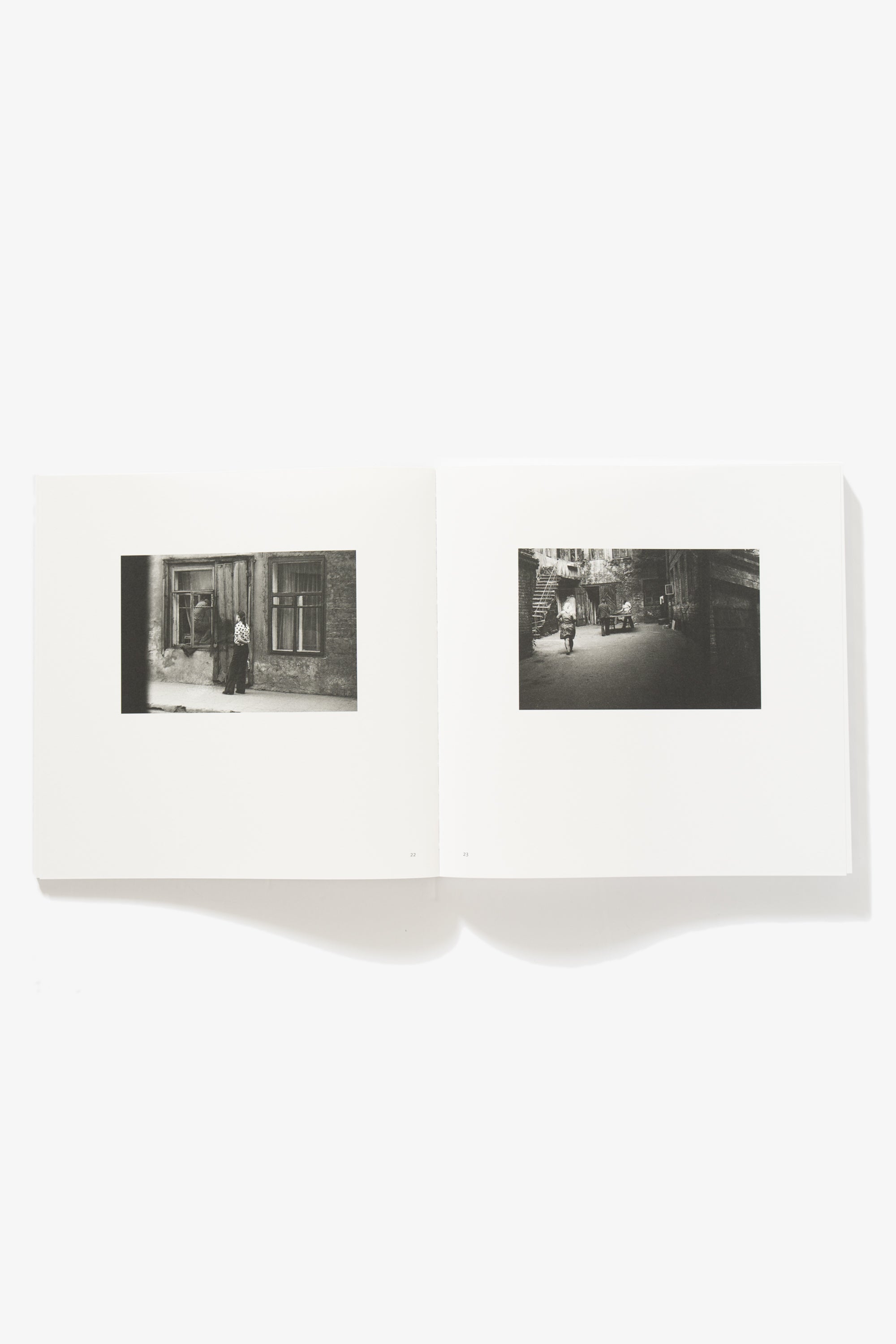 Spread of Grey But Not Grey. Verso shows a woman standing in front of an entrance, shot in middle distance, turned away from camera. Recto shows the inner courtyard of a residence complex, two figures standing aside a table, a woman walking into the courtyard. 