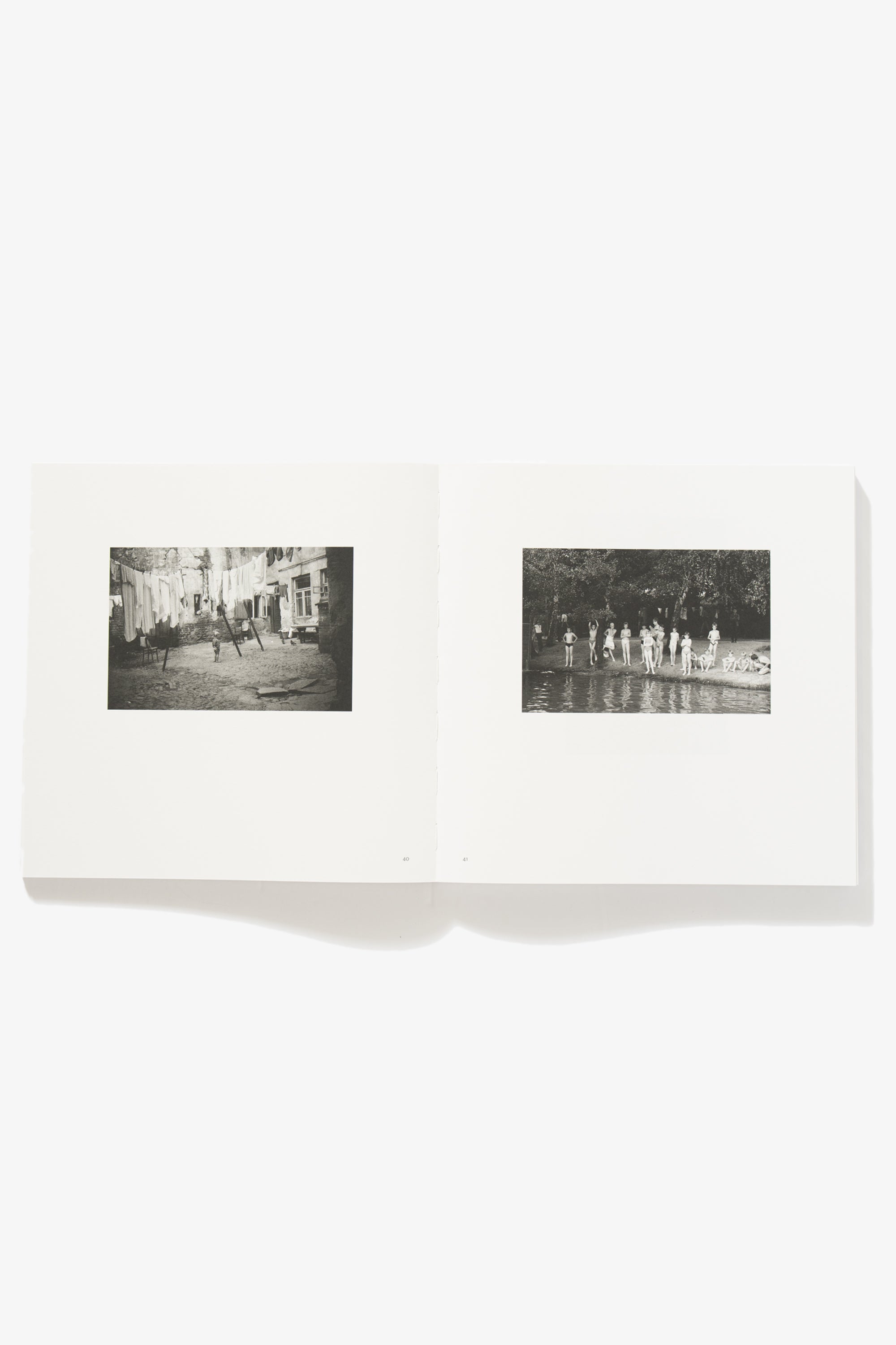 Spread of Grey But Not Grey. Verso shows a courtyard with laundry sagging on lines hung in a brick courtyard, a part of the ground damaged. A young child stands beneath them. Recto depicts youths standing on the shore of a lake, preparing to swim.
