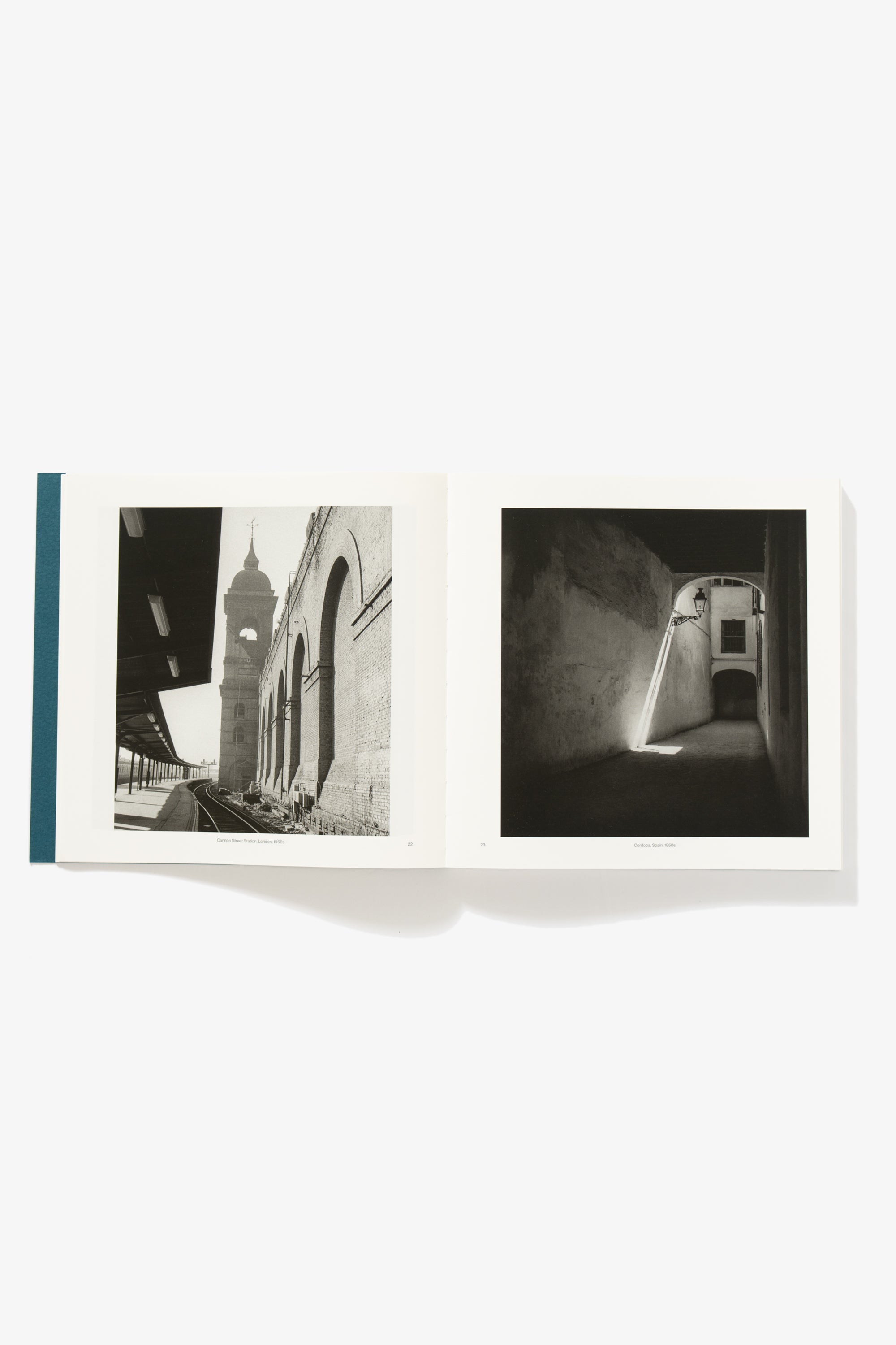 Spread of A World Observed. Verso depicts the length of a metro train track, a clock tower in the distance, a retaining wall with arcs sculpted in relief running parallel to the camera’s perspective. gaze. Recto depicts a darkened covered pedestrian pathway, a street light extending from the wall, with two parallel beams of light extending behind it to the ground.