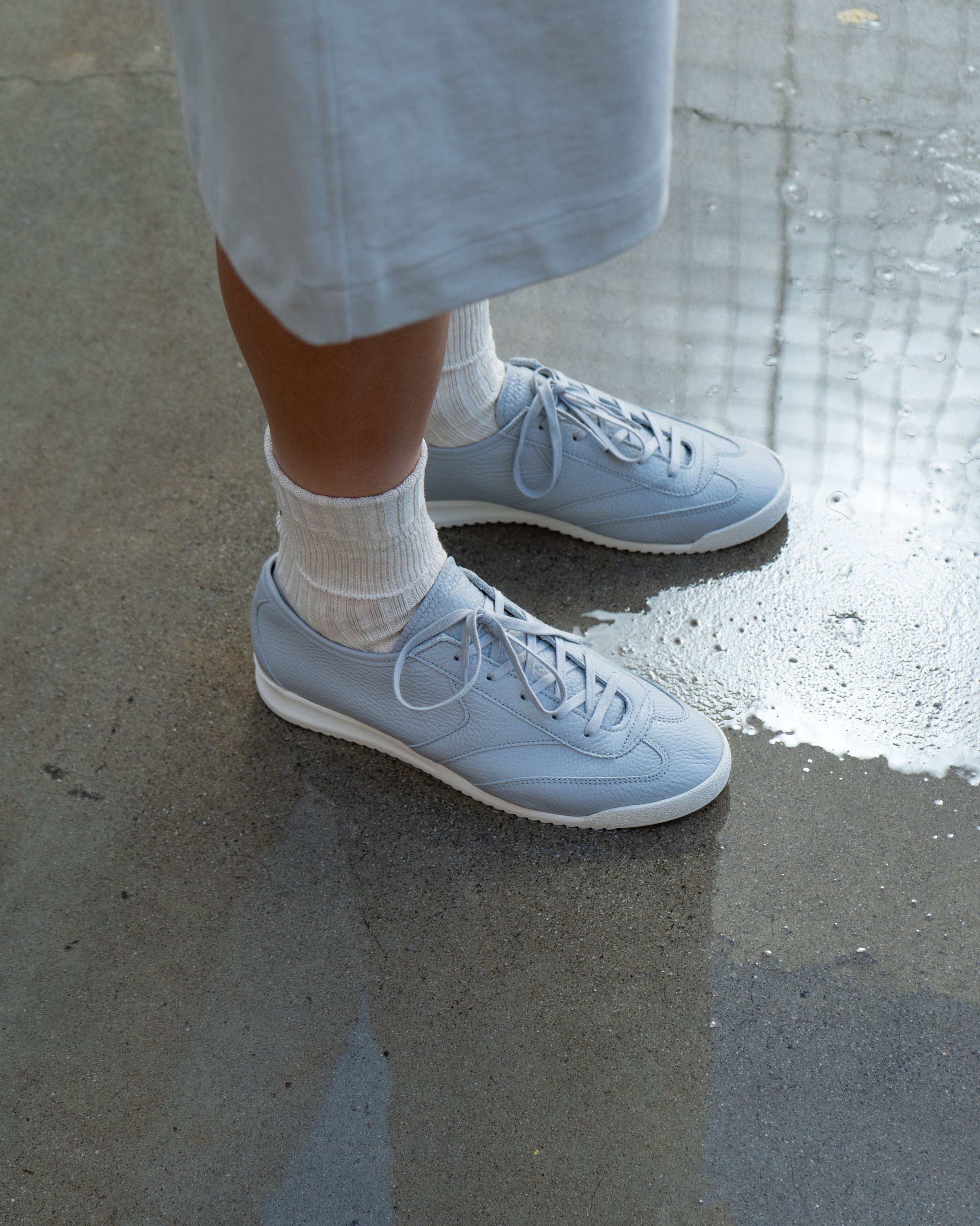 Close focus of LW1070 German Military Sneakers in Aluminum on model, the trunk of LW1150 Jersey Shorts in Swiss natural extending from the top frame. Feet posed on wet ground.
