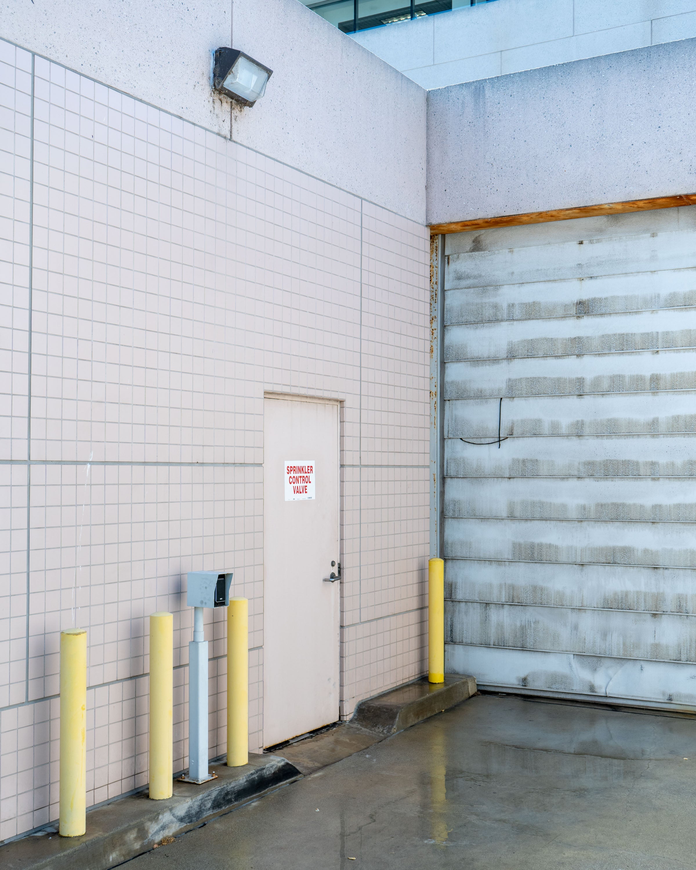 Still image of a loading bay, tiled background on the left with a back entrance door; the interleaved panels of the steel garage door is partially in view