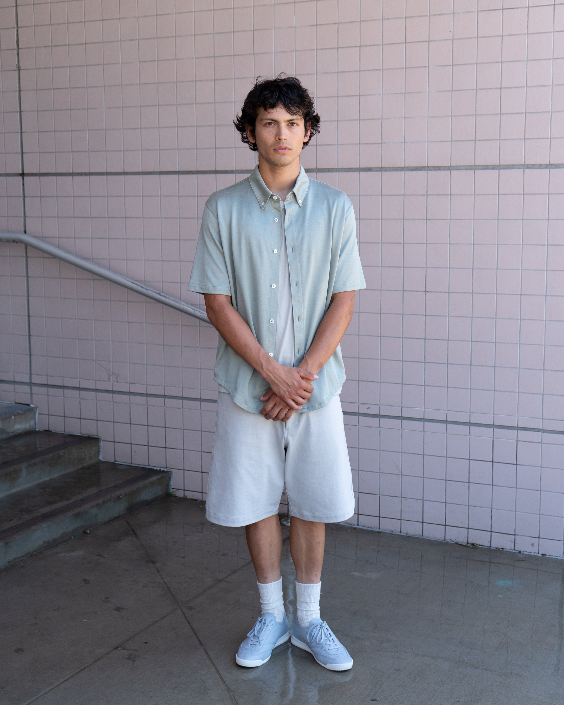 Model wears LW423CR SS CR Button Down in Swiss natural, LW1150 Jersey Short in Swiss Natural, and LW1070 German Military Sneaker in Aluminum a few steps away from a low staircase against a tiled background