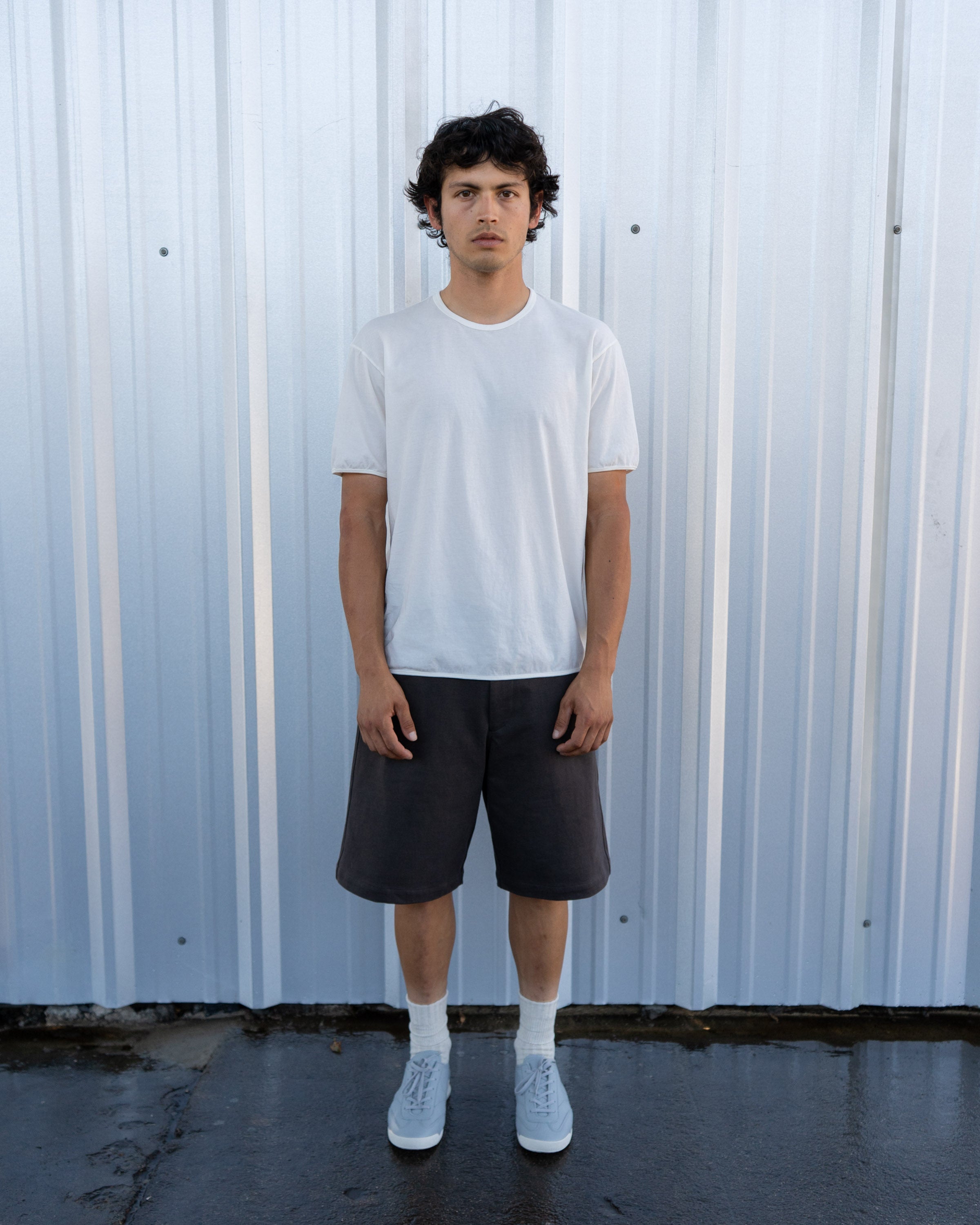 Model wears a LW110 Dry Lite T-shirt in White, LW1150 Jersey Short in Black Pine, and LW1070 German Military Trainer against a corrugated metal background on wet concrete ground