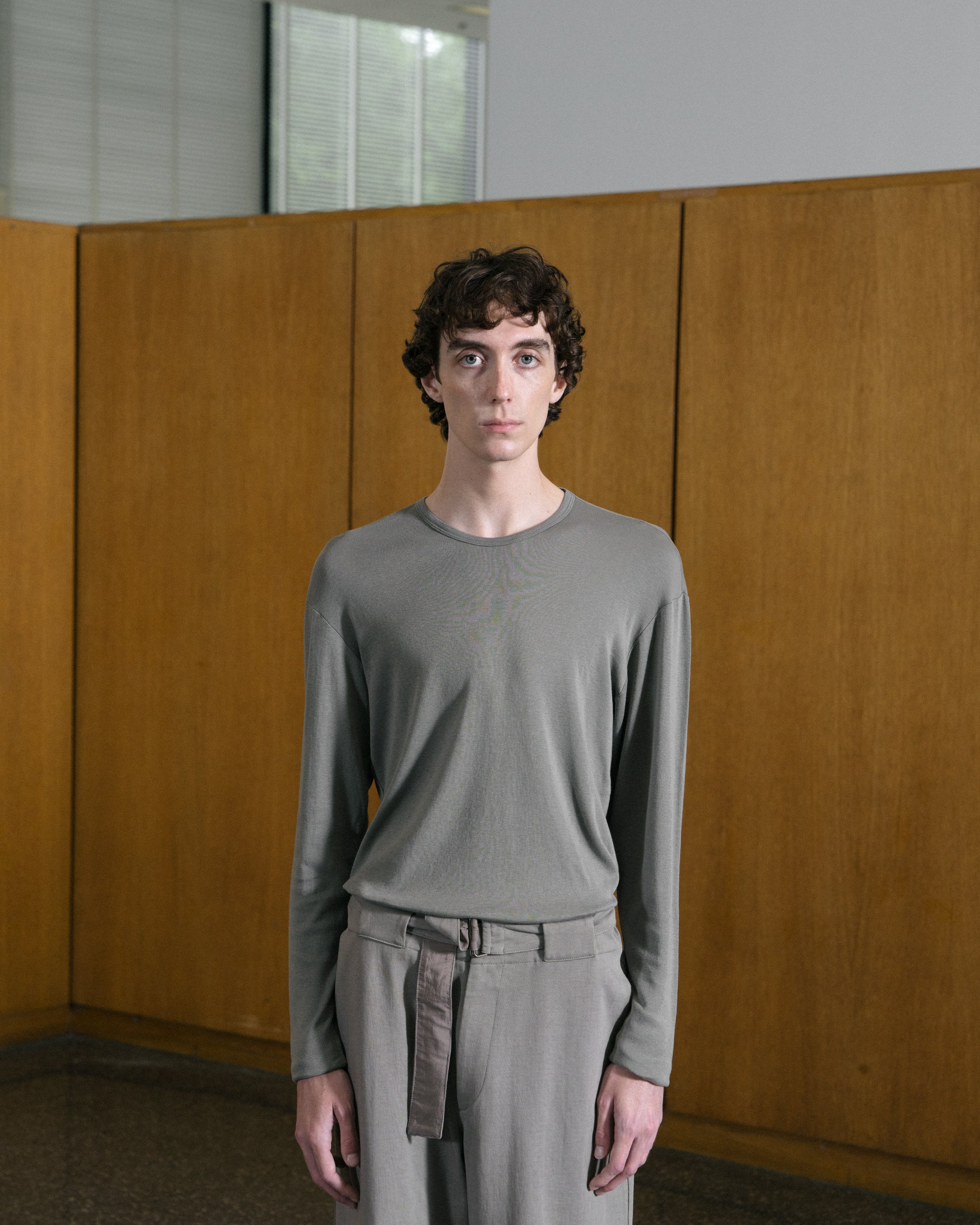 Model stands against a tan paneled wall, wearing the LW201 Carlen L/S T-shirt in Anvil Grey, and Pigment Chalk trousers
