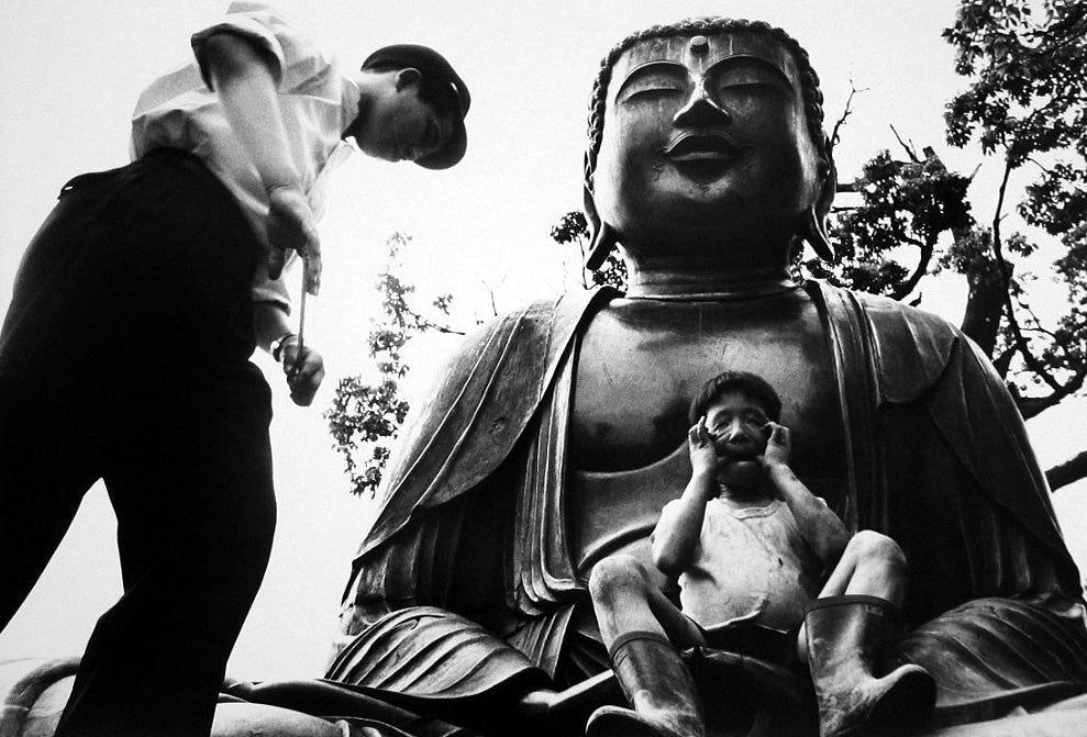 Large Buddha statue with a person and child sitting on its lap, with another person standing nearby.
