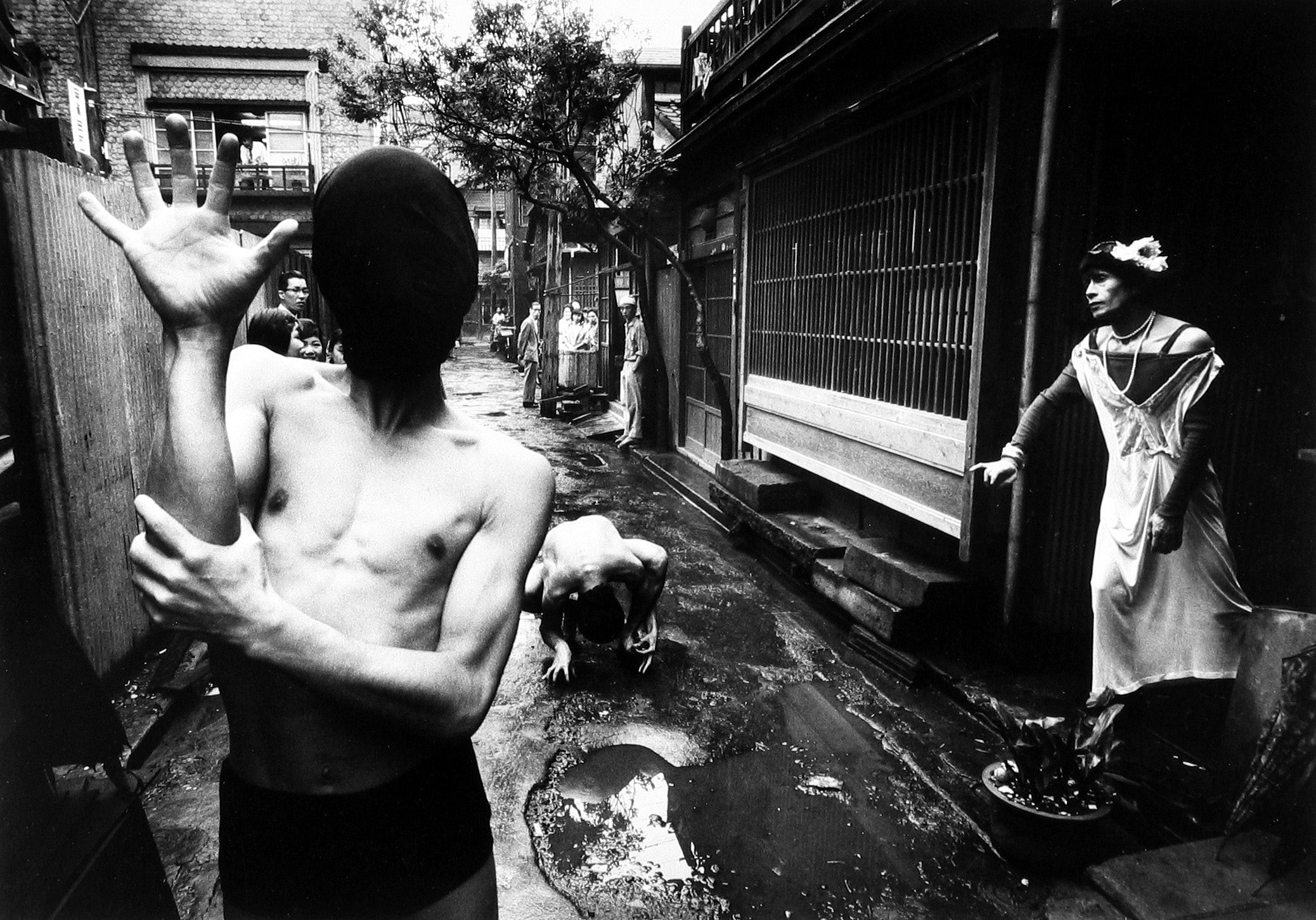 Still from William Klein's 1961. Butoh dancers pose in an alleyway. The figure in the foreground obscures his face with an opaque hood. 