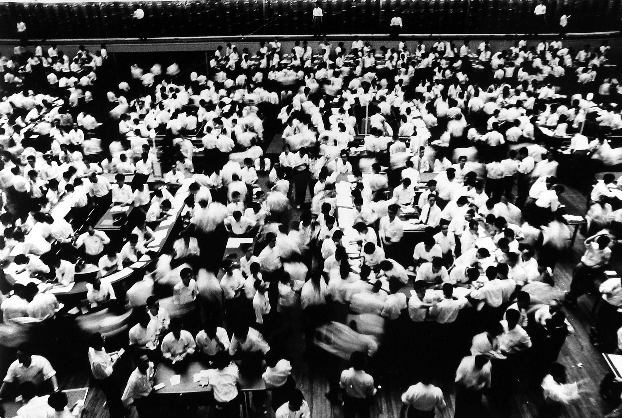 Still from William Klein's Tokyo 1961. A large crowd of people rush about in a large room, resembling the floor of a market or exchange hall. 