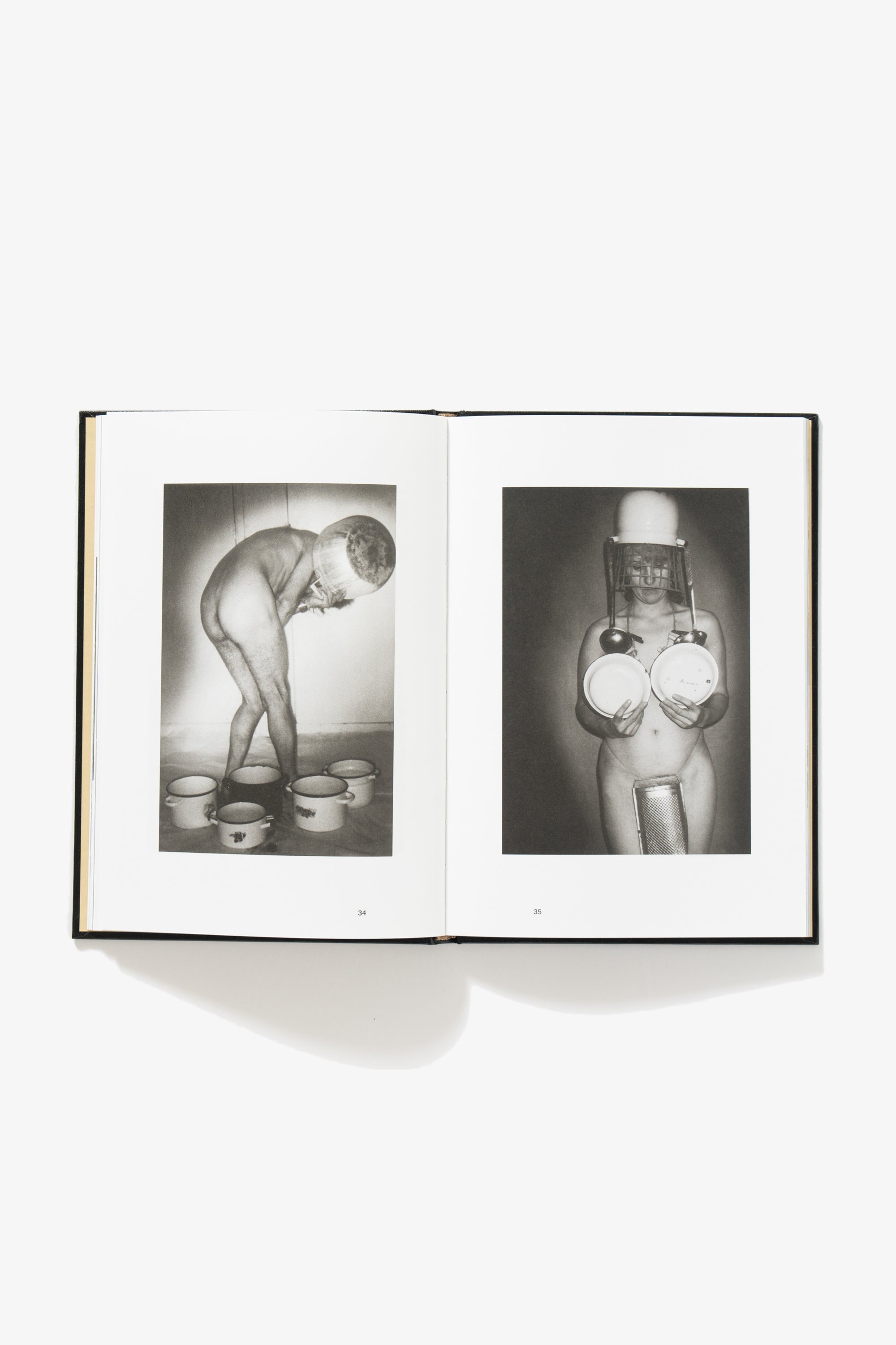 Spread of NAKED. Verso shows a figure of indeterminate sex turned away from the camera, hunched over, obscuring their face with one of the pots strewn about their feet. Recto shows a female-appearing person covering chest and genitals with pots and kitchen equipment, a mixer tipped over their head with ladles hanging from the sides in imitation of long hair and earrings.