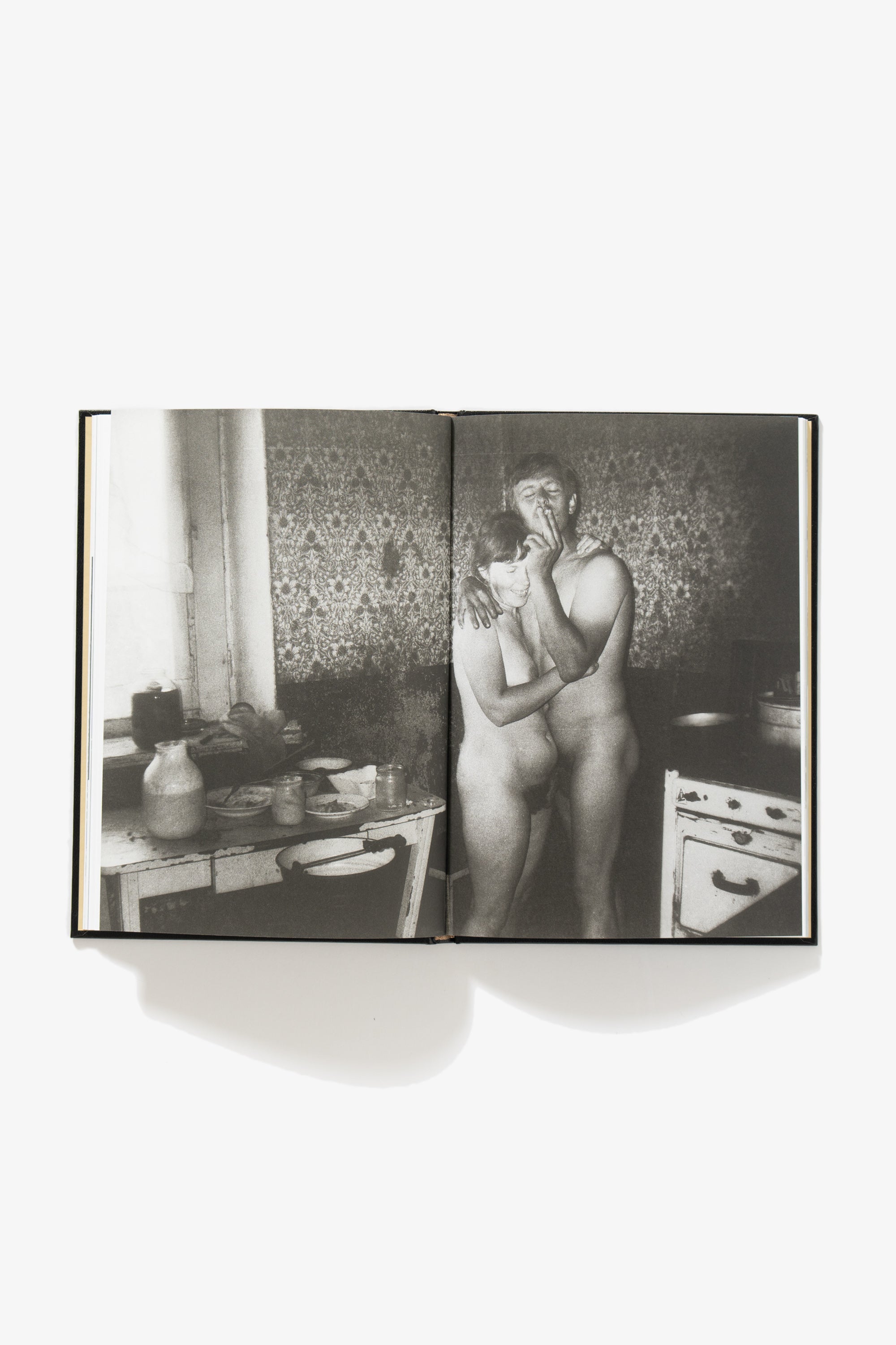 Spread of NAKED. One image occupies both pages. Image depicts a nude couple in a room. The wife is embraced by the husband, smoking a cigarette. A kitchen table occupies the verso.