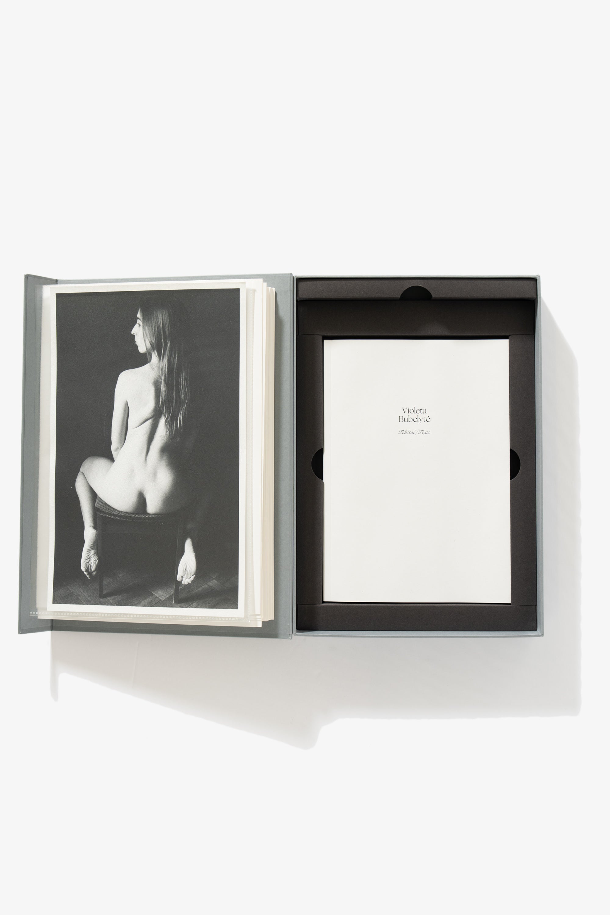A full-length image of the collection opened. The left-side shows a woman sitting on a chair, her back toward the camera, her nude buttocks visible. The right side is the cover of a collection of texts, titled “Violeta Bubelyte.” 