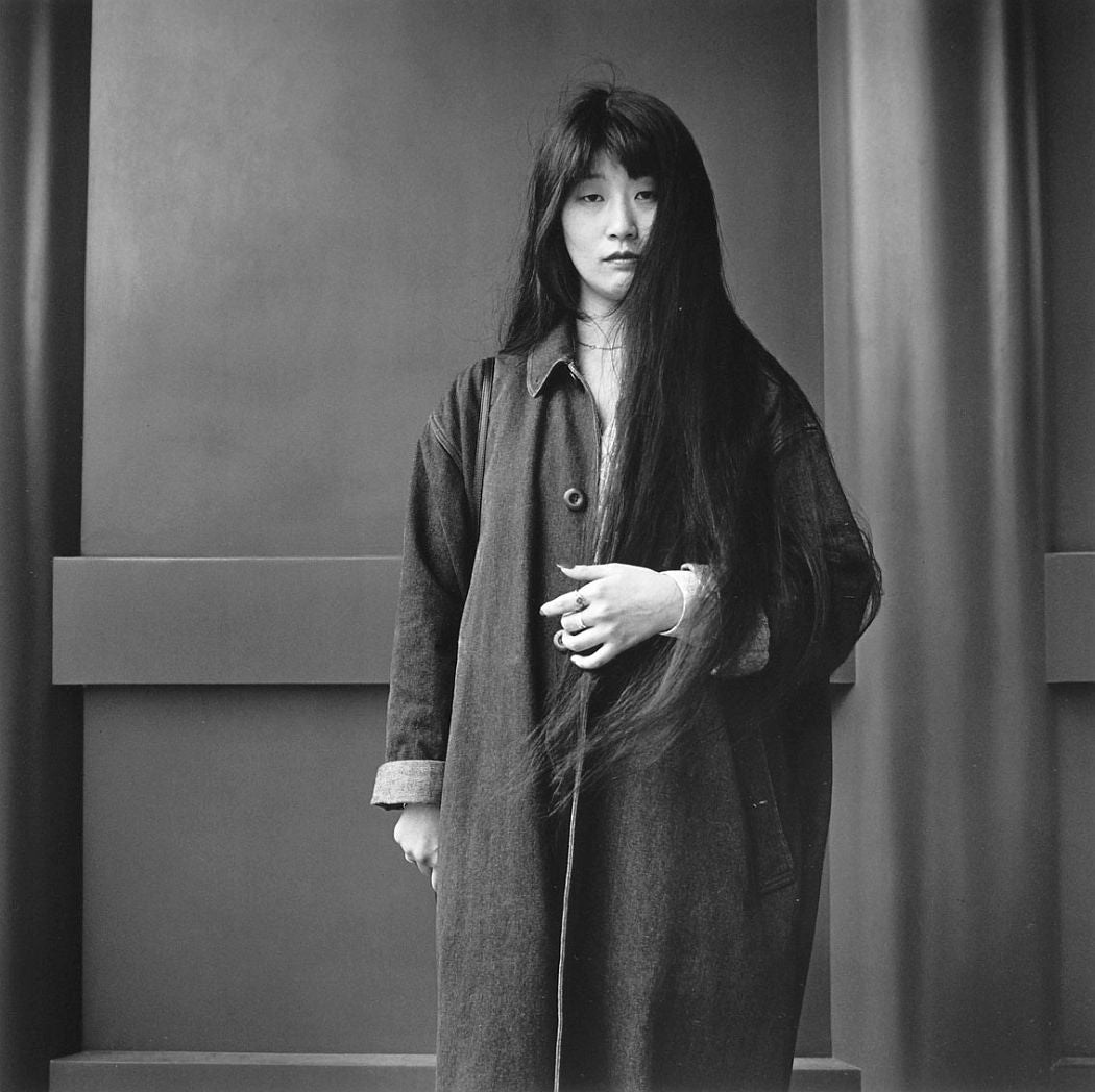 Photograph from Hiroh Kikai's "Asakusa Portraits." Portrait of a woman wearing a long coat, her long hair stretching to her waist, against a plain background.
